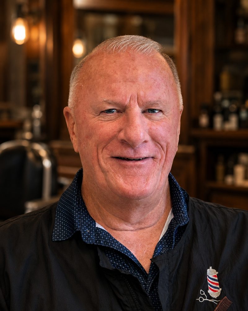 Perry, barber, smiling in the shop wearing a dark barber jacket with a barber-pole pin.